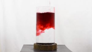 An image of a glass of water on a black stand against a white background. Red liquid is being poured into the full glass.