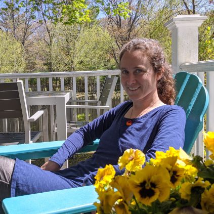 Photo of Jennie. She is on a teal reclining Adirondack chair. Her light curly hair is pulled back into a low ponytail. She is wearing a dark blue dress. Yellow pansies are in the foreground.