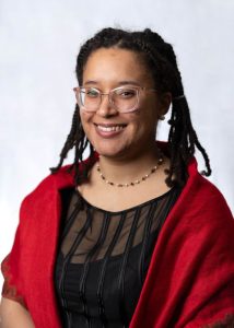 Katarina smiling at the camera in a red shawl and black dress, wearing a necklace and glasses. Her hair is in braids and pulled neatly to the side