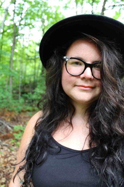 Rebecca looks into the camera, wearing a black dress, a black hat, and glasses