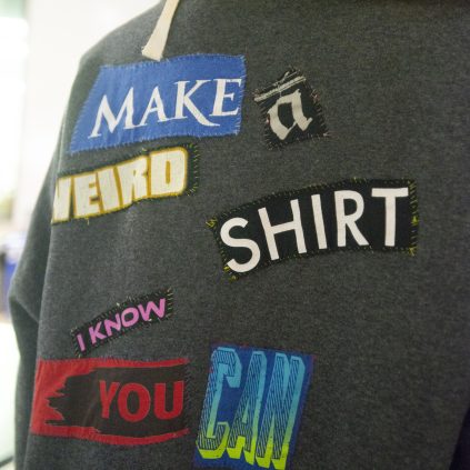 A photograph of the back of a sweater onto which patches from t-shirts have been sewn by hand. The patches each have one word printed in a variety of fonts and colors, and spell out the sentence "Make a weird shirt, I know you can."