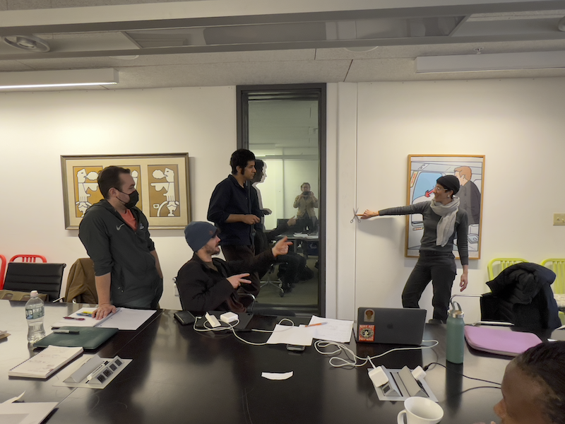 Students in a meeting room, one student is holding an open pair of scissors against the wall with one finger while others look on.