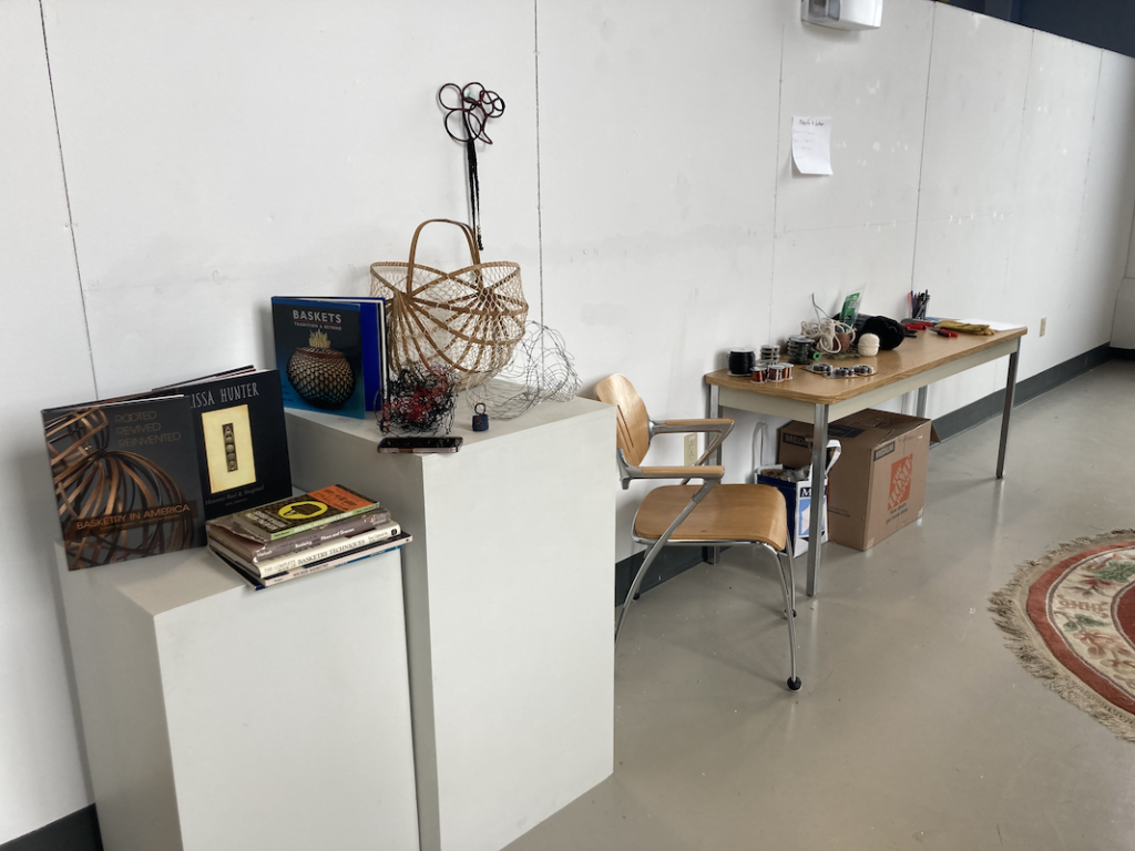 Two white pedestals against a white wall, on top of which are art books and wire sculptures.