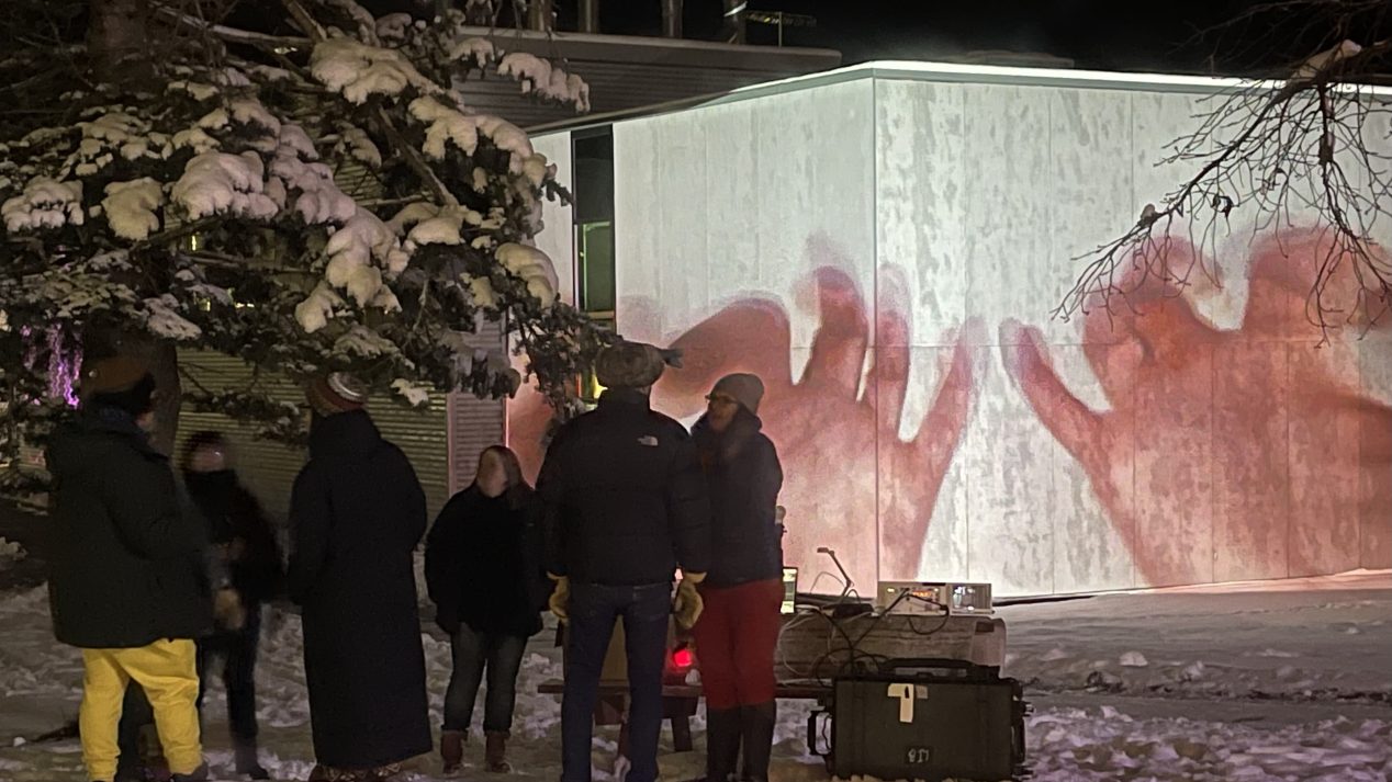 at night, a collection of people gather outside in the quad covered in snow to view projection mapping projects that cover the outside of the building
