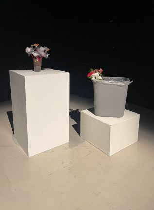 Jenna Davenport's work with flowers made from recycled materials sitting in a McDonalds on one pedestal, and a bouquet of real flowers placed in a trash bin on the pedestal to the right