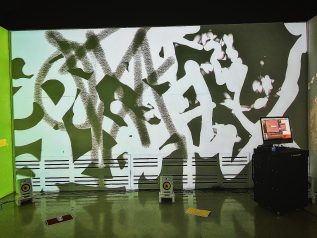 black and white, graffiti-like visuals projected onto a wall, directional speakers sit on the floor and a kiosk cart with a computer running the program sits to the right.