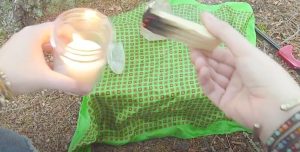 close up of two hands in front of a green cloth over grass, right hand holds a small burning item, left hand holds a candle lit