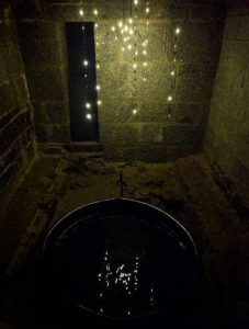 in a darkly lit space with sting lights hanging in the background over brick, there is a metal bucket filled with a liquid reflecting the light