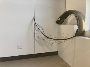 a temporary installation made out of found scrap metal which appears to curl inwards like a spiral, installed in a corner of the room against a white wall