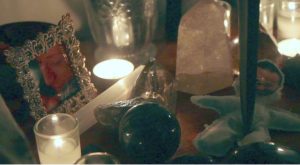 a collection of trinkets gather on a table to make what looks to be a shrine, there are candles and picture frames and a voodoo doll with someones photo added as the face, there is a dagger pushed into the chest of the voodoo doll
