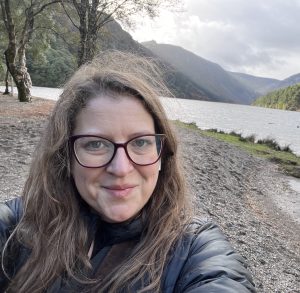 Woman with shoulder length light brown taking a selfie in front of a river and mountains
