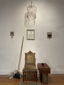 an old chair sits in the center of frame with a broom to its left and frames hung on the wall behind it, an old suitcase is propped up to the right of the chair like a side table, a bird cage hangs above with a light bulb like a chandelier