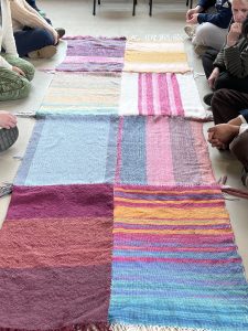 a long rectangular woven tapestry with bright colors like pink and blue and orange in different directions, people sitting around each at their own spot
