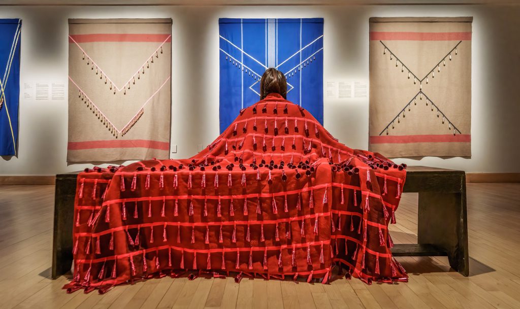 Woman sitting on bench with her back to us, in red textile draped over her back and the bench, person appears to be looking out at three more textiles hung on a wall